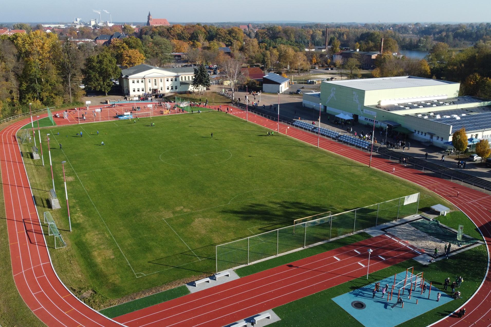 Luftaufnahme eines Sportgeländes mit Fußballplatz, Laufbahn und Freizeitanlage. Bäume und Gebäude im Hintergrund.
